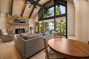 A modern living room with a stone fireplace, a sofa set, a dining table, and large windows offering a view of trees and a building. - Sunset Spectacular | Big Bear Lake Views, Walk to Meadow Park, Ping Pong & Fenced Yard (Big Bear Lake)