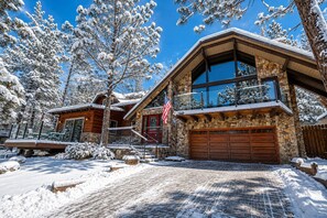 A snow-covered lodge with a stone facade, a large garage, and a balcony overlooking the snowy landscape. - Sunset Spectacular | Big Bear Lake Views, Walk to Meadow Park, Ping Pong & Fenced Yard (Big Bear Lake)