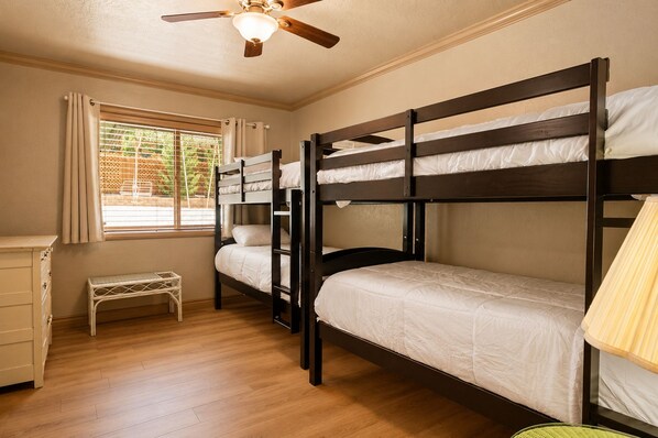 Une chambre avec un lit superposé, un ventilateur de plafond, une fenêtre avec des rideaux, un petit banc et une table de chevet.