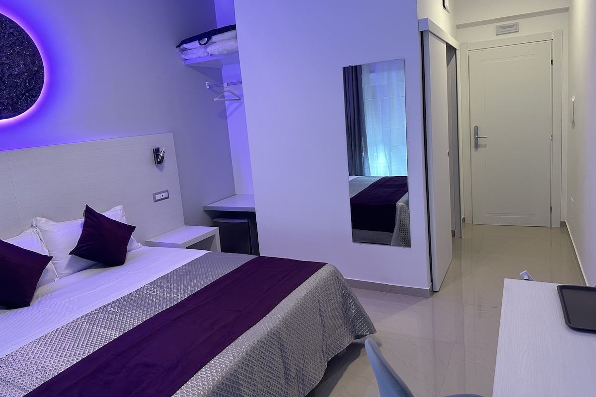 A modern hotel room with a bed, bedside tables, a mirror, and a door.