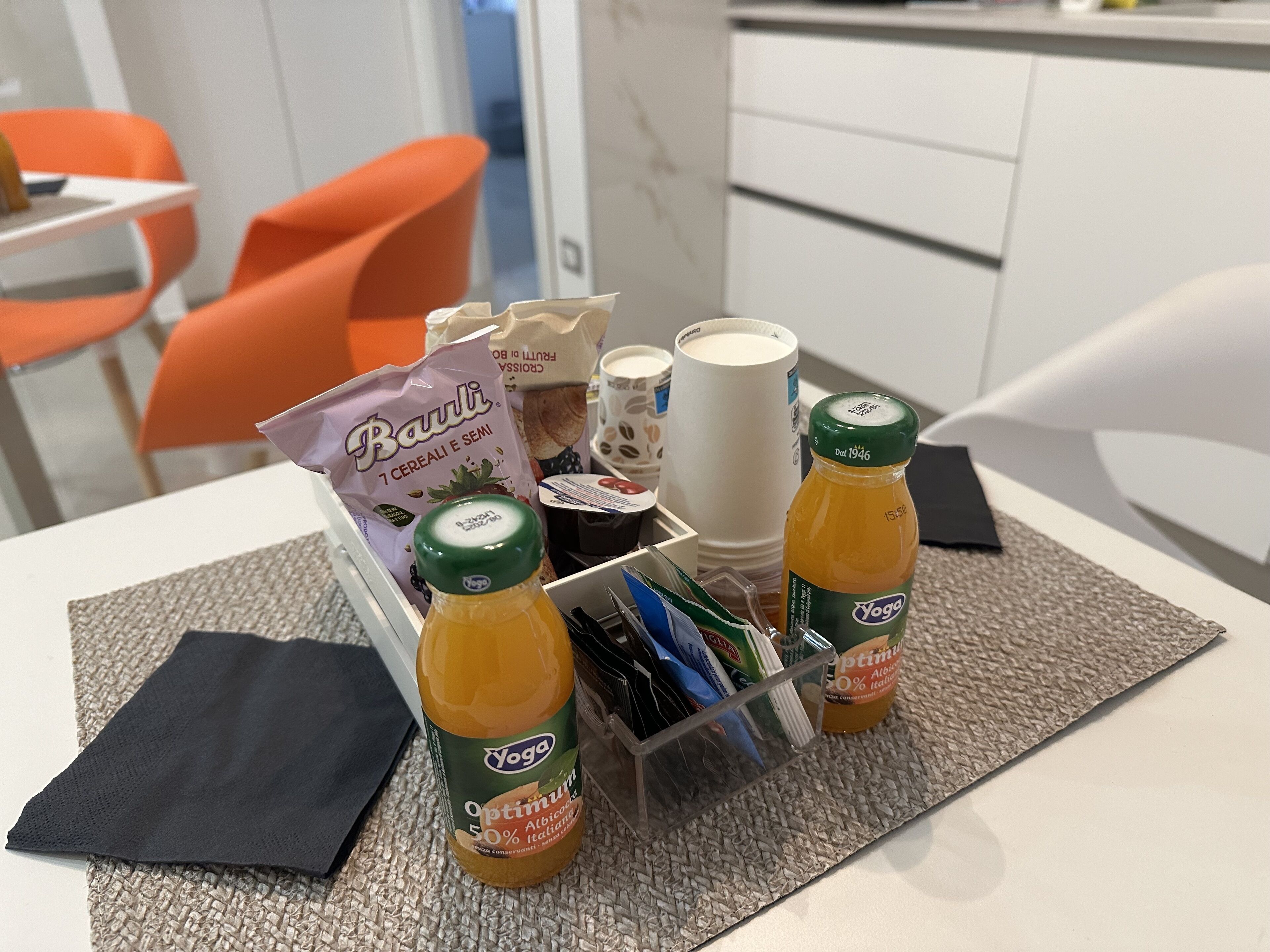 Ein Frühstücksaufbau mit einer Müslischachtel, einer Saftflasche, einer Tasse und einem Becher auf einem Tisch.