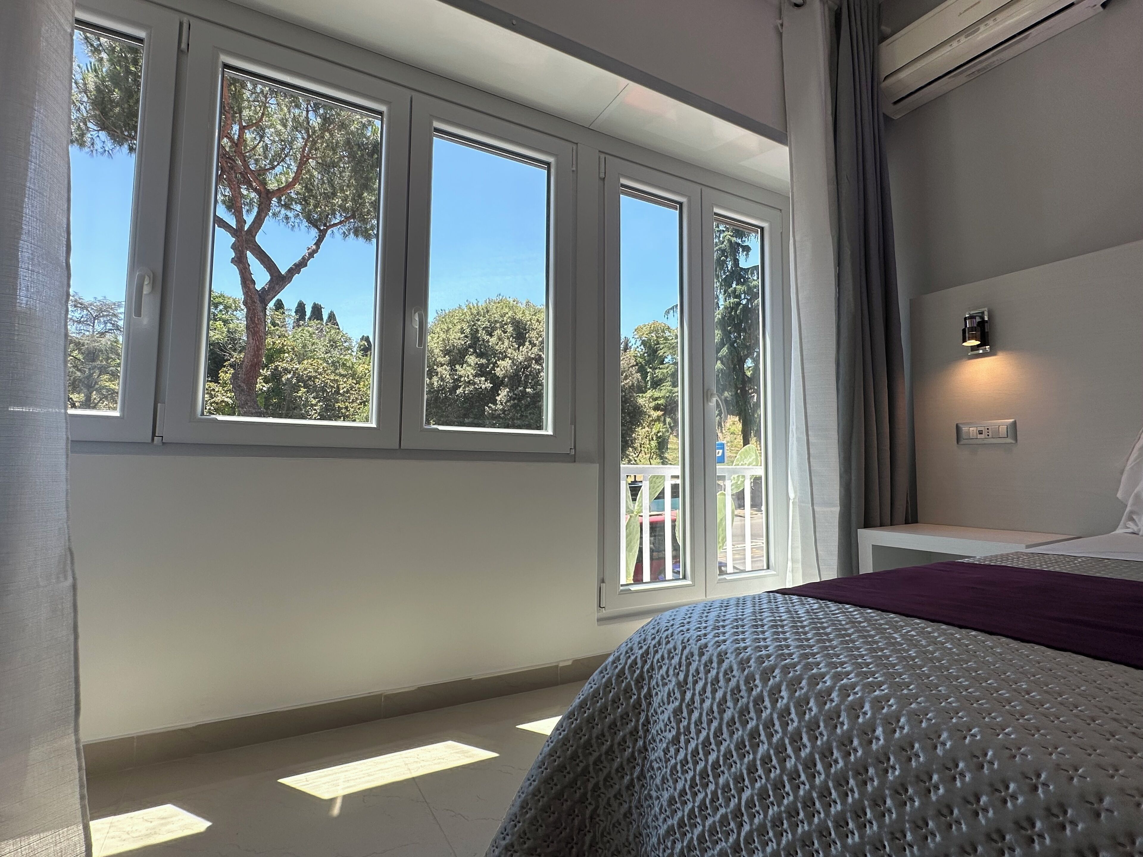 A bedroom with a large window, a bed, a bedside table, and a wall-mounted light fixture.