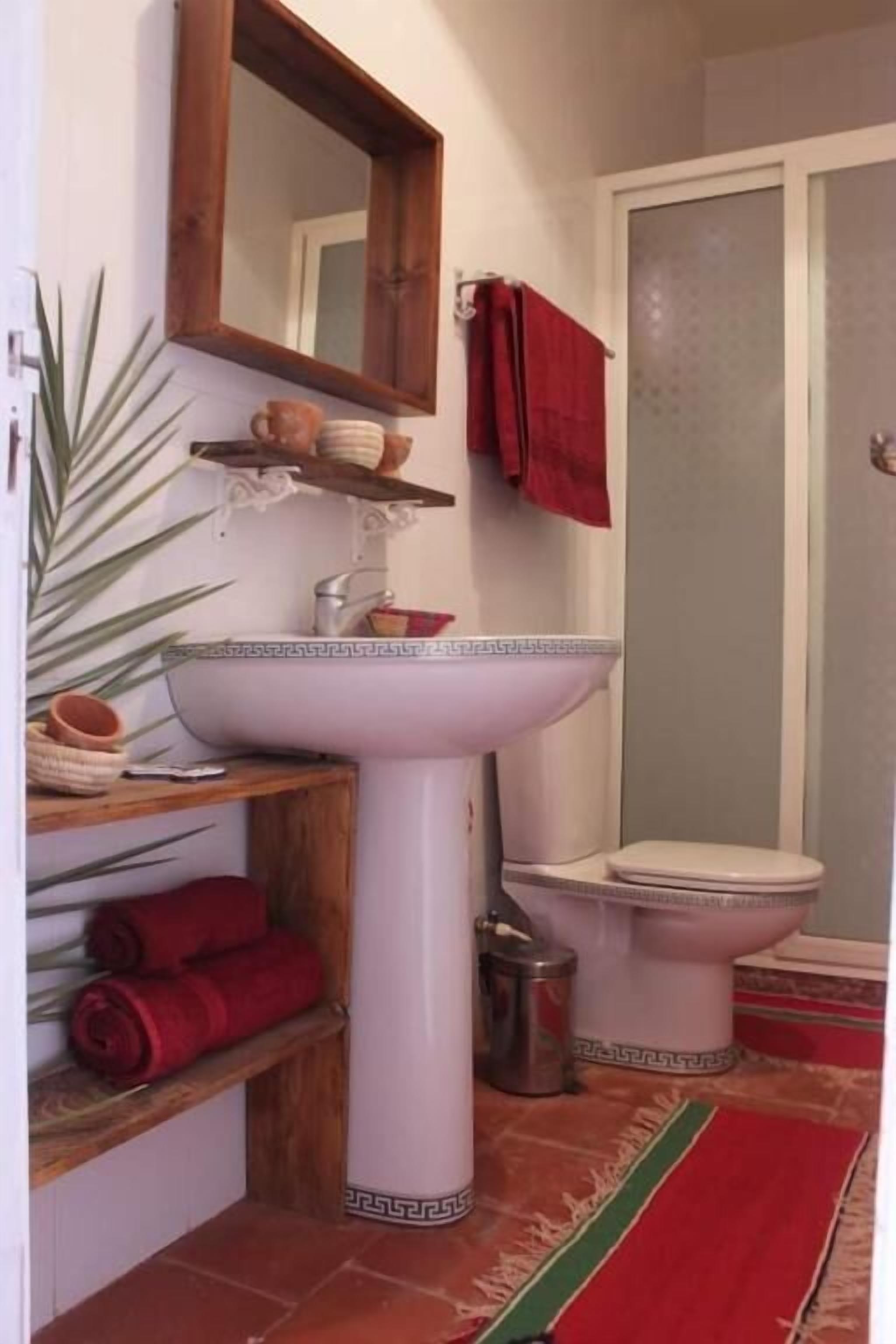 A bathroom with a white pedestal sink, a toilet, a mirror, and a red towel on a shelf.