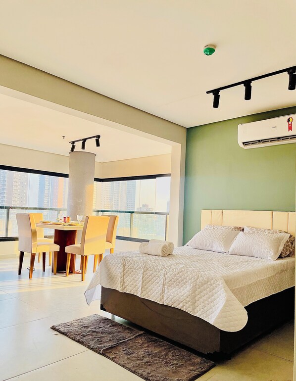 Ein modernes Hotelzimmer mit einem großen Bett, einem Essbereich mit Tisch und Stühlen sowie Blick auf die Stadt durch große Fenster.