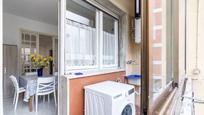 Ein Waschbereich mit Waschmaschine, einem Fenster mit Spitzenvorhängen, einem Tisch mit Stühlen und einer Vase mit gelben Blumen.