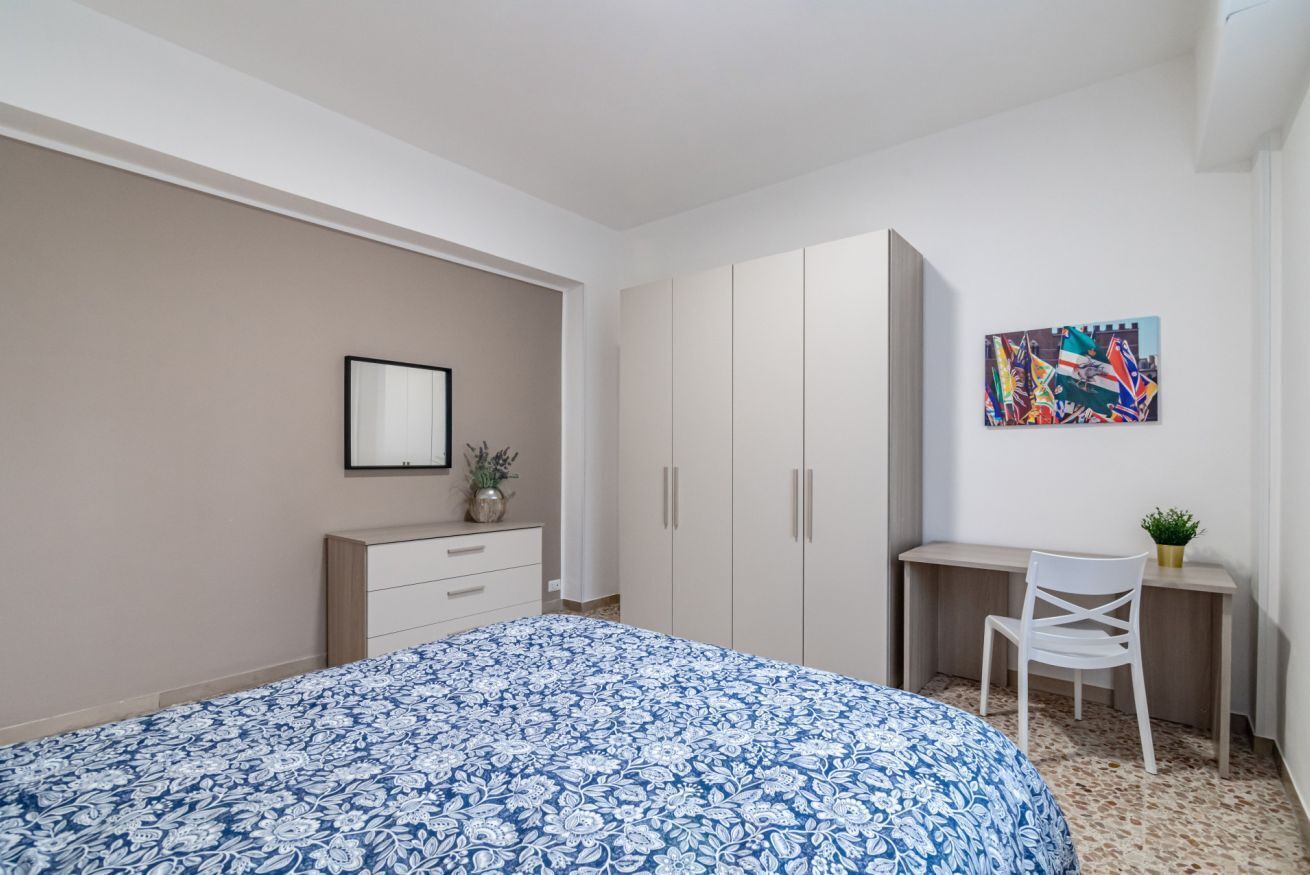 A bedroom with a bed, a desk, a chair, a wardrobe, and a framed picture on the wall.