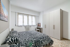 A bedroom with a bed, a window with curtains, a radiator, a desk, and a wardrobe.
