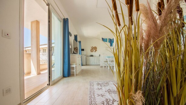 Habitación de hotel con puerta de vidrio corredera que da a un balcón, zona de cocina y sala de estar con una planta grande.