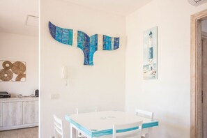 A dining area with a white table and chairs, a wall-mounted artwork resembling a whale, and a kitchen area with a marble countertop.
