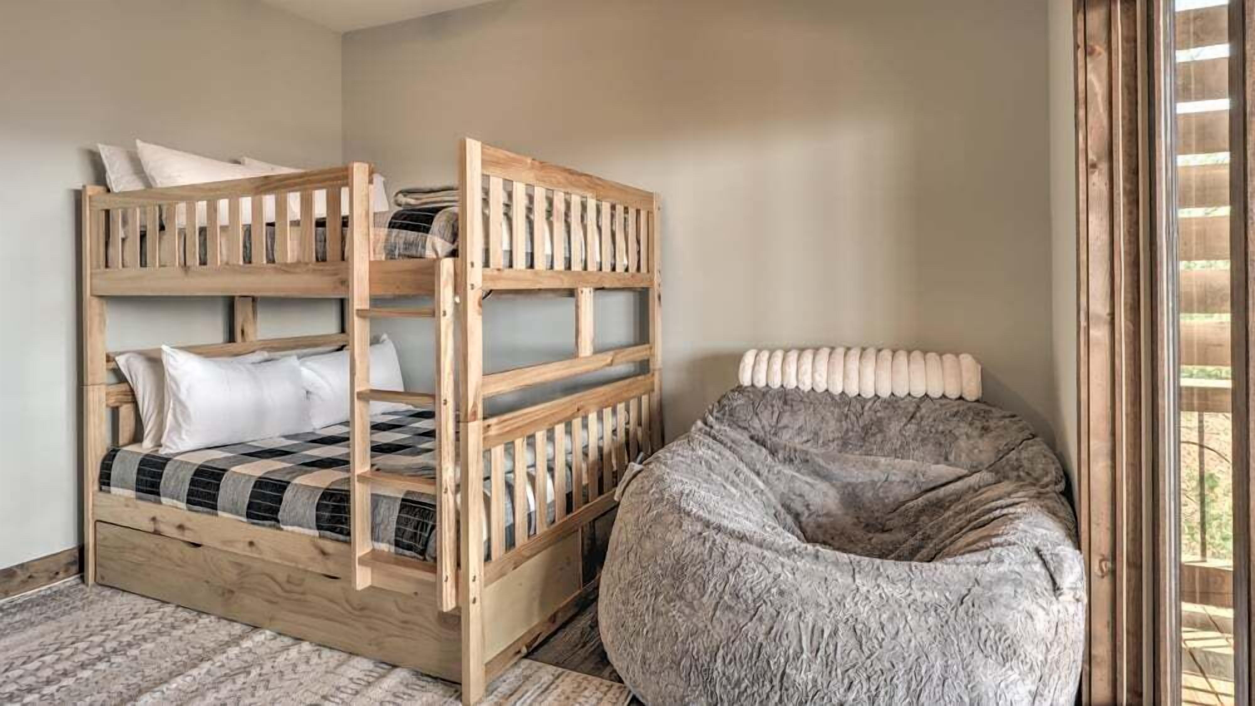 A bunk bed with a checkered blanket and pillows, a round bean bag chair, and a window with blinds.