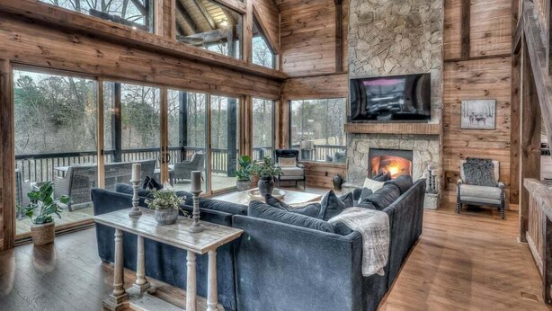 A cozy living room with a stone fireplace, comfortable seating, and large windows offering a view of the outdoors.