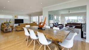 A spacious living room with a large wooden dining table, white chairs, and a modern orange sculpture. There are multiple seating areas with sofas and a television. - Beautiful Vibe Energy Escape Lux w Bonuses Phillip Island 100m to nature inlet (Rhyll)