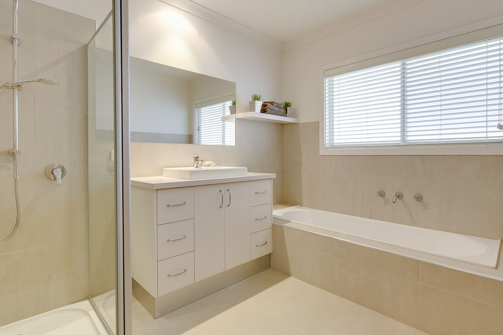Un baño moderno con cabina de ducha de vidrio, un lavamanos blanco con espejo arriba, una bañera y una ventana con persianas.