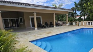 A swimming pool with lounge chairs, a covered patio, and palm trees.