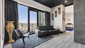 A modern living room with a black leather sofa, a large floor-to-ceiling window with a city view, a tall decorative vase, and a blue patterned rug. - The VIBE PALMS PLACE PENTHOUSE  (Las Vegas)
