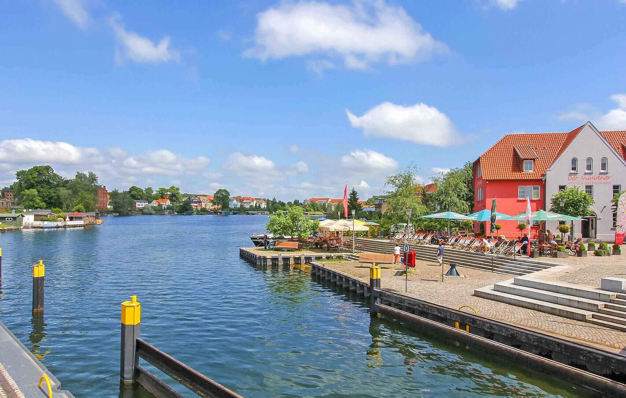 Une vue sur le fleuve, avec un quai, un bâtiment rouge portant l’inscription « Hôtel et restaurant », et une rangée de chaises longues.