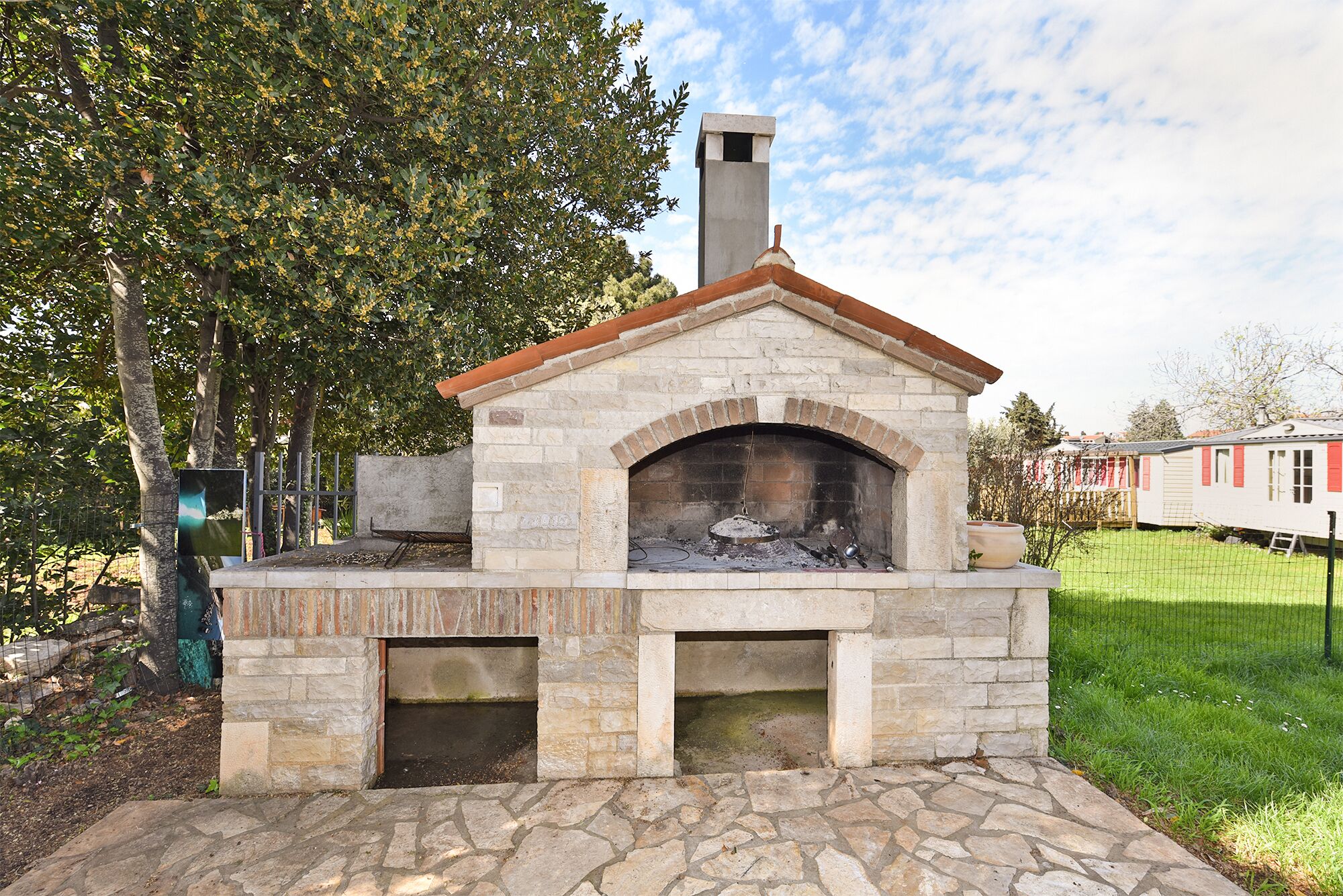A stone-built outdoor pizza oven with a chimney, surrounded by a stone-paved area and a grassy field.
