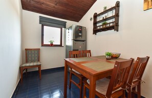 A dining area with a wooden table and chairs, a refrigerator, a window with a plant, and a shelf with glassware.
