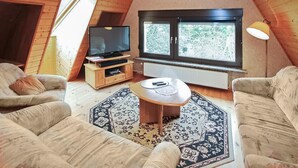 Ein gemütliches Wohnzimmer mit Fernseher, zwei Sofas, einem Couchtisch und einem großen Fenster mit Blick auf Bäume.
