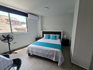 Une chambre d’hôtel équipée d’un lit, d’un bureau, d’une chaise, d’un ventilateur sur pied et d’une fenêtre avec un appareil de climatisation.
