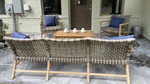 Un patio con mobiliario de mimbre y una mesa de madera, una puerta marrón y un muro de ladrillo.
