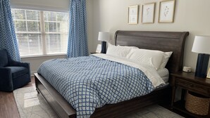 Un dormitorio con una cama de madera, ropa de cama azul estampada, un sillón azul, una mesita de noche con lámpara y una ventana con persianas.