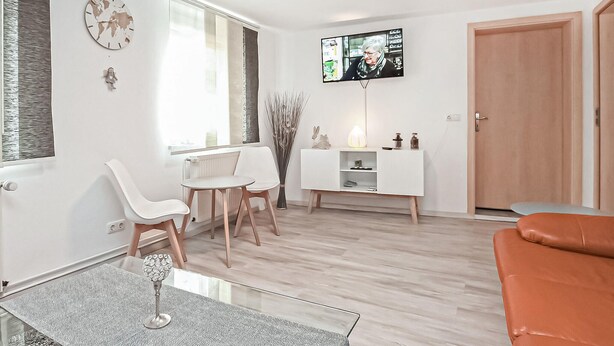 A modern living room with a white sofa, a dining table with chairs, a TV mounted on the wall, and a wooden door. - Ferienhaus Röbel/Müritz (Röbel/Müritz)