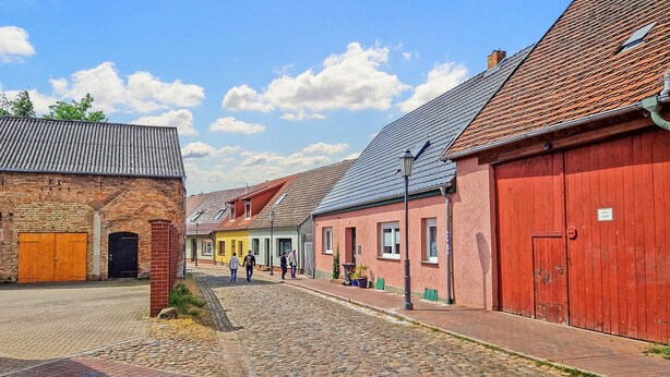 Una calle empedrada bordeada por edificios históricos, entre ellos un edificio de ladrillo con una puerta de madera grande y un edificio rojo con un letrero que indica restaurante.