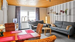 Una sala de estar moderna con techo de madera, un sofá gris, una mesa de comedor con individuales rojos y una ventana con cortinas.