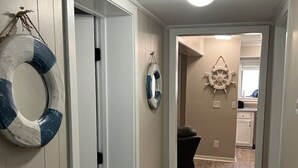 Un couloir doté d’un plancher en bois, de murs blancs et d’une porte donnant sur une pièce. On y voit deux bouées de sauvetage accrochées au mur, ainsi qu’une décoration représentant un gouvernail de navire.
