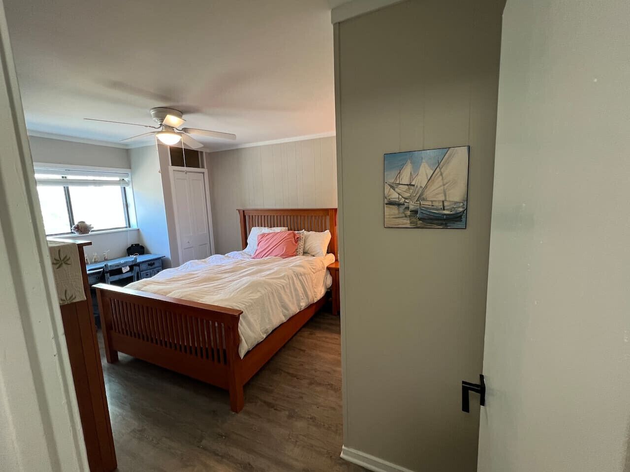 A bedroom with a wooden bed, a ceiling fan, a window with blinds, and a painting on the wall.