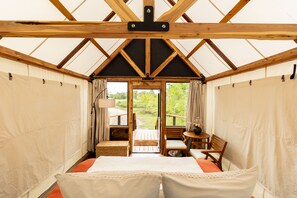 A cozy, well-lit tent-style room with a bed, a small table, and a chair, featuring a wooden ceiling and large windows.
