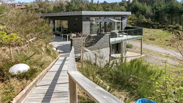 Une maison moderne dotée d’une terrasse en bois qui mène à une entrée en verre, le tout entouré d’une végétation naturelle.