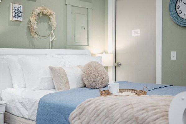 Een net opgemaakt bed met een blauwe deken, witte kussens en een zachte plaid. Een klein mandje met een kop en decoratief object staat op het bed.