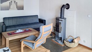 Uma sala de estar aconchegante com um sofá, uma cadeira de madeira, uma mesinha com um controle remoto, uma lareira e uma cesta de vime.