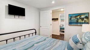 A bedroom with a bed, a TV mounted on the wall, a door leading to another room, and a painting of palm trees on the wall. - "Recuperation Cottage" offers gulf views and is steps to the beach! Pet-friendly, boat parking (Port Saint Joe)