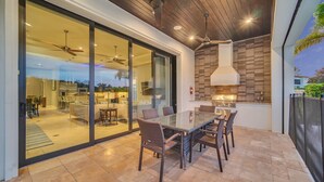 Un espace repas extérieur avec une table en verre et des chaises en osier, un ventilateur de plafond et une vue sur l’intérieur à travers des portes vitrées.