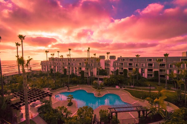 A resort with a pool, palm trees, and a view of the ocean at sunset.