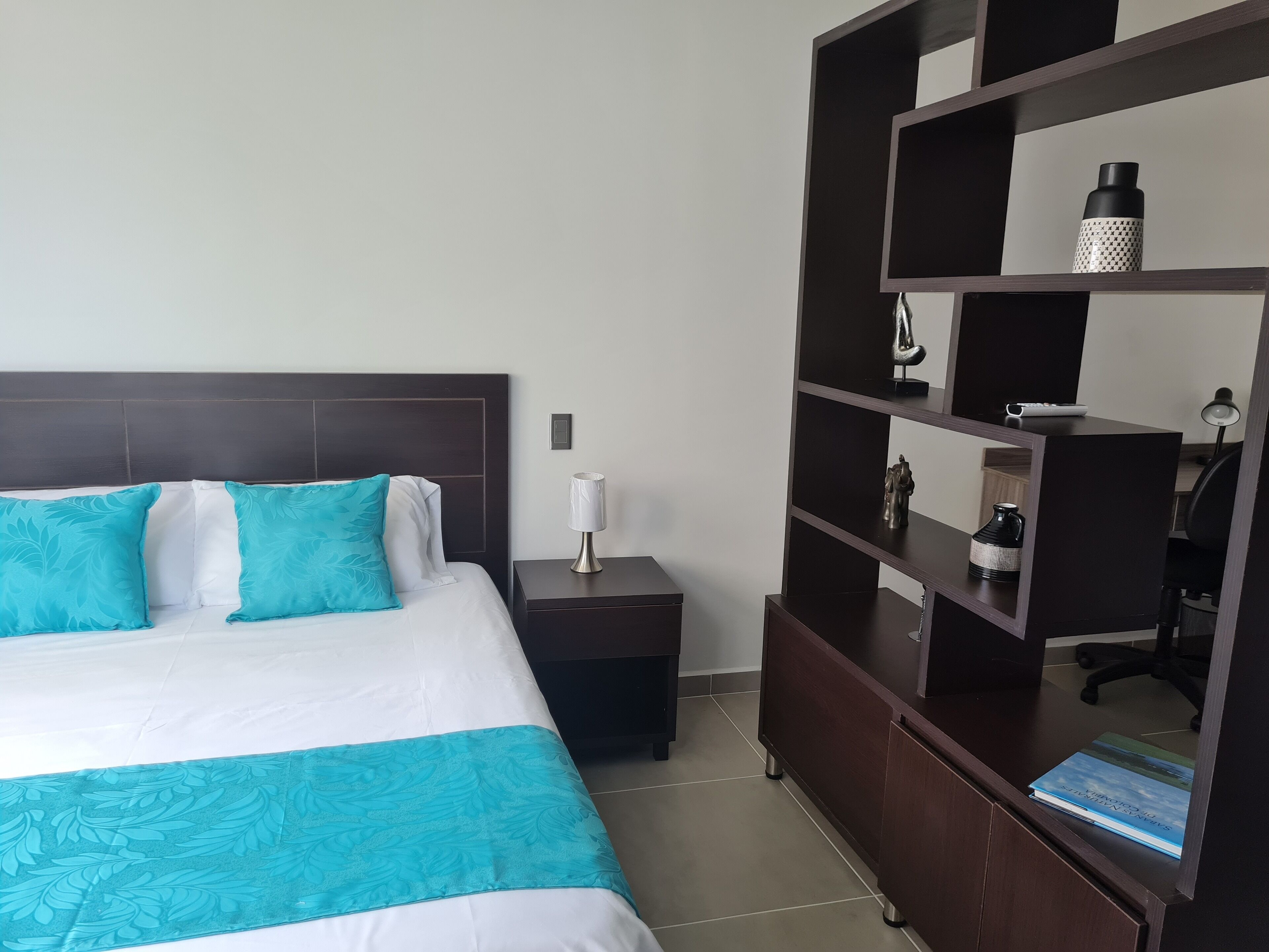 A hotel room with a bed, a nightstand, a lamp, a shelf with decorative items, and a book.