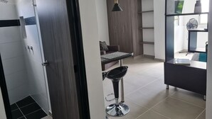 Un baño moderno con ducha, inodoro y lavamanos. Cuenta con una pequeña zona de estar con una mesa y una silla. La habitación tiene un ventanal grande y un ventilador de techo.
