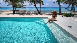 A clear blue pool with a woman sitting on the edge, surrounded by palm trees and beach chairs.