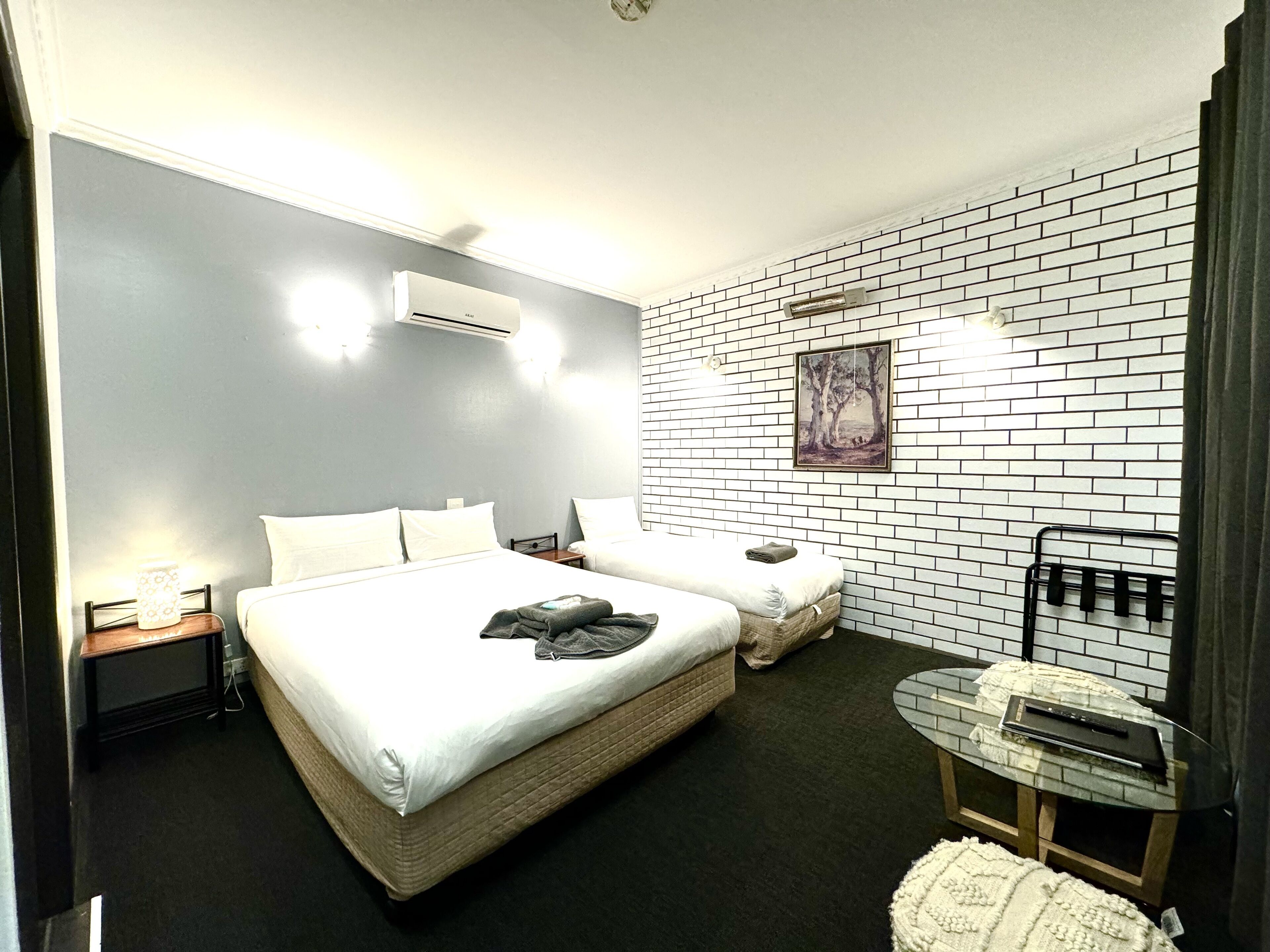A hotel room with a large bed, a smaller bed, a round table, a chair, a wall-mounted TV, and a painting.