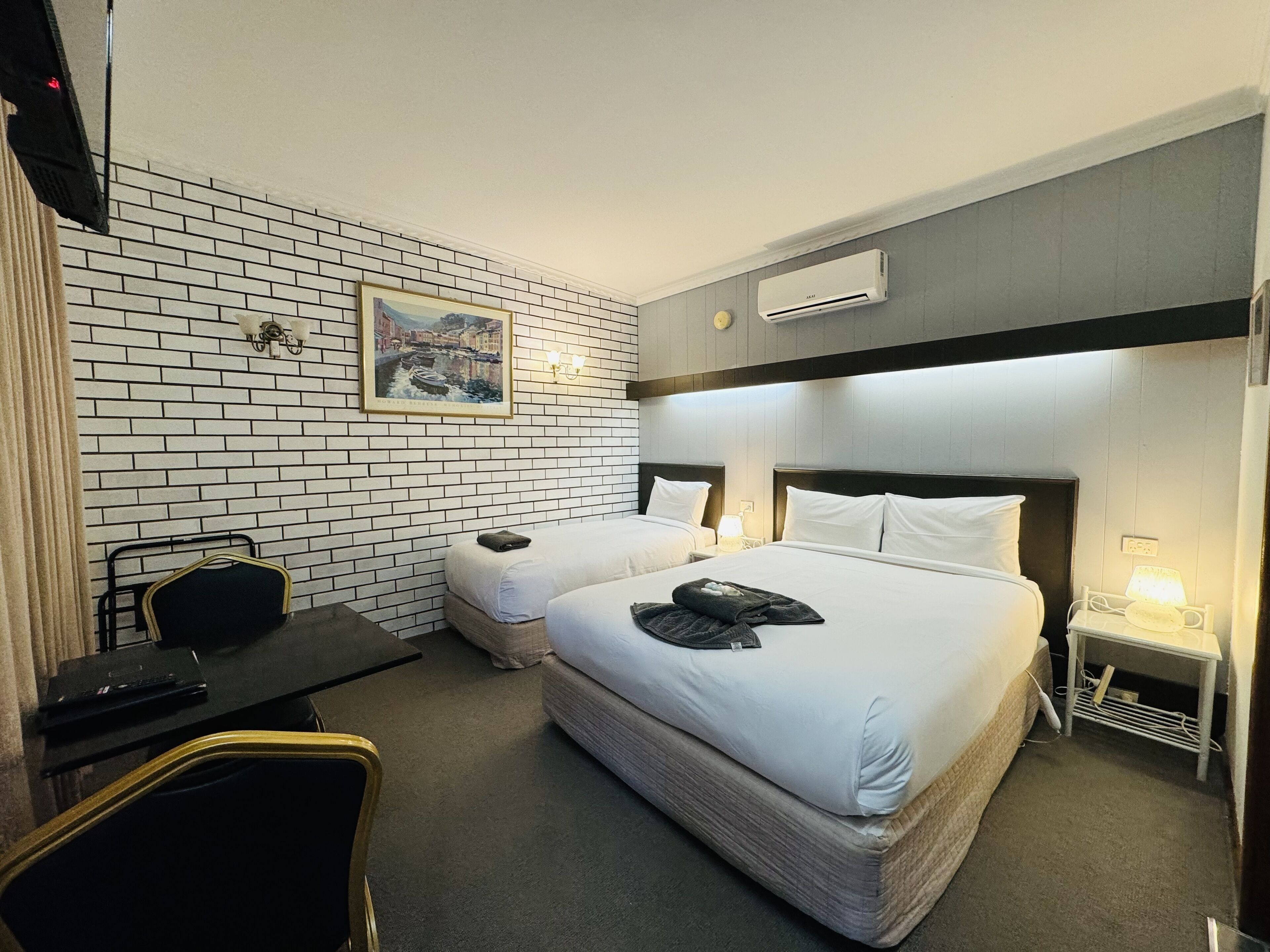 A hotel room with two beds, a desk, a chair, a painting on the wall, and an air conditioning unit.