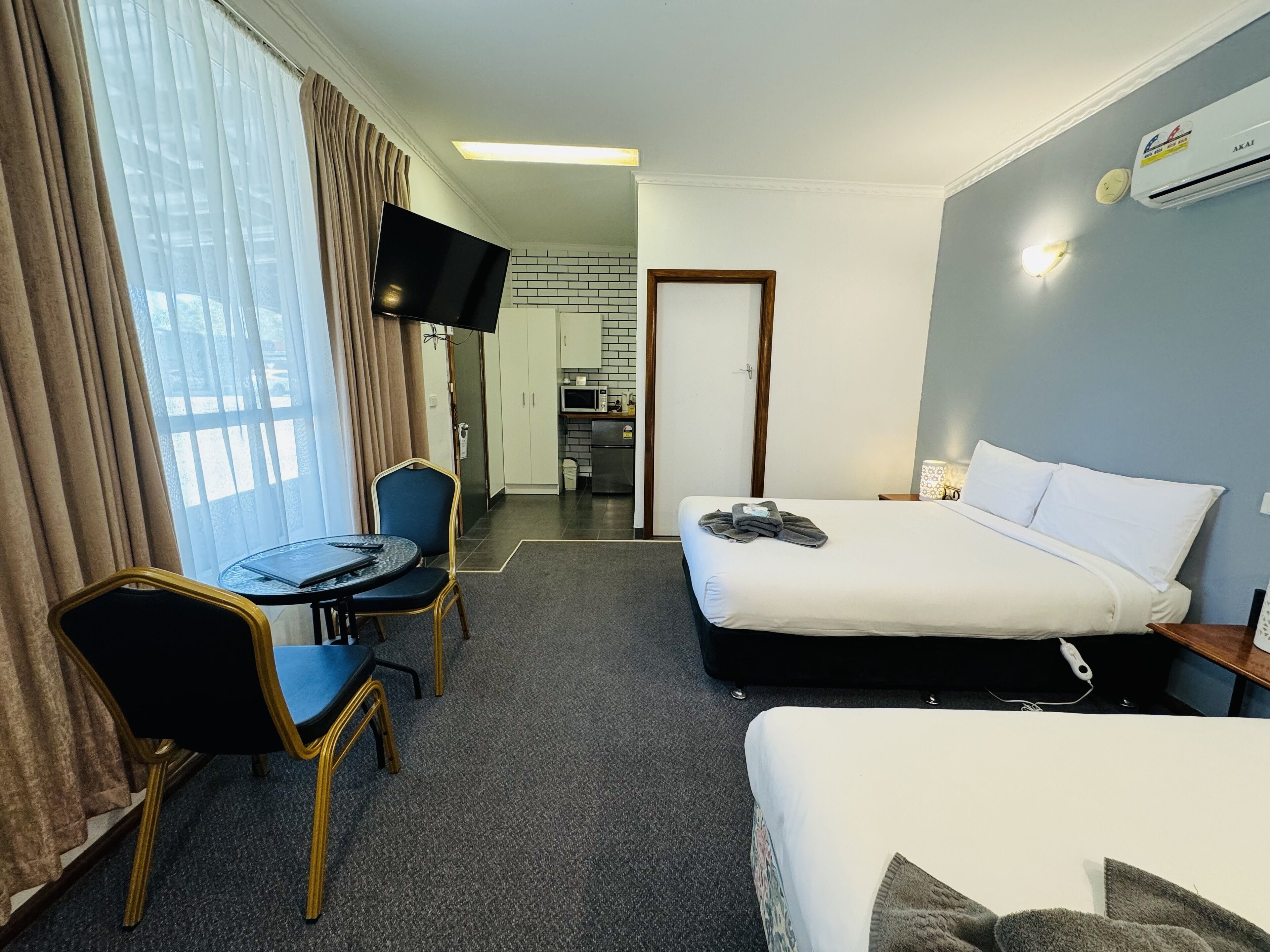 A hotel room with a bed, a desk with a chair, a television, and a kitchenette in the background.