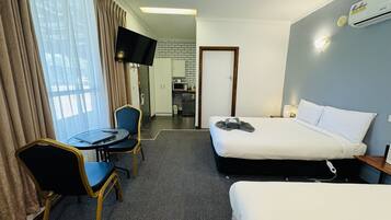 A hotel room with a bed, a desk with a chair, a television, and a kitchenette in the background.