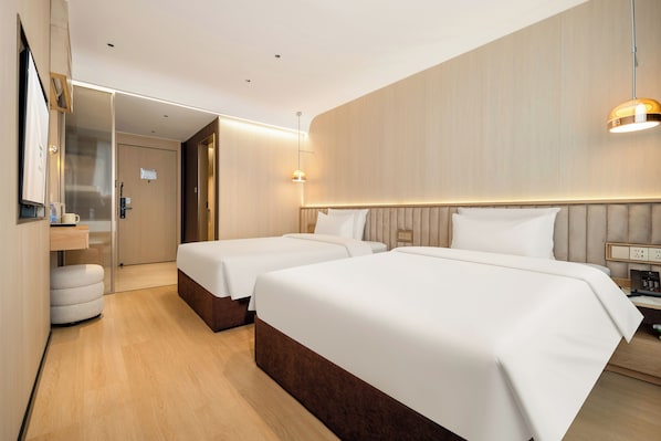 Habitación de hotel con dos camas, suelo de madera, un minibar y una mesita de noche con lámpara.