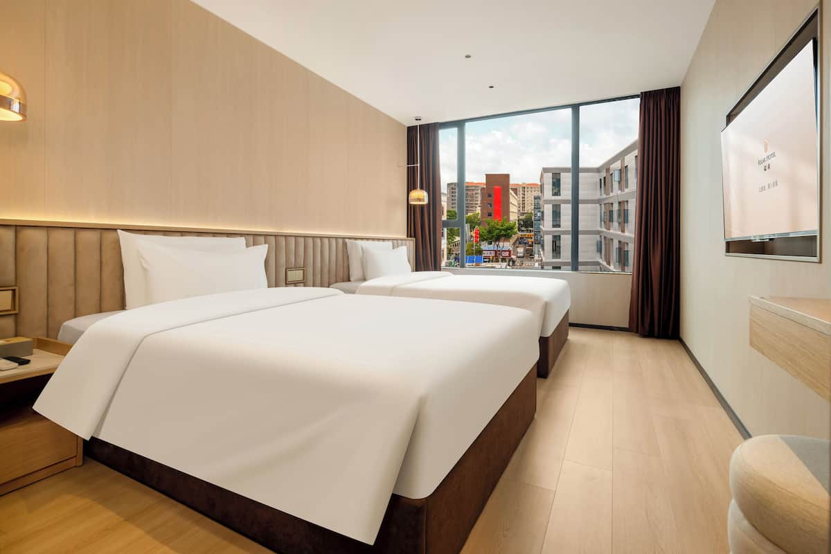 A hotel room with two beds, a large window with a city view, and a flat-screen TV on the wall.