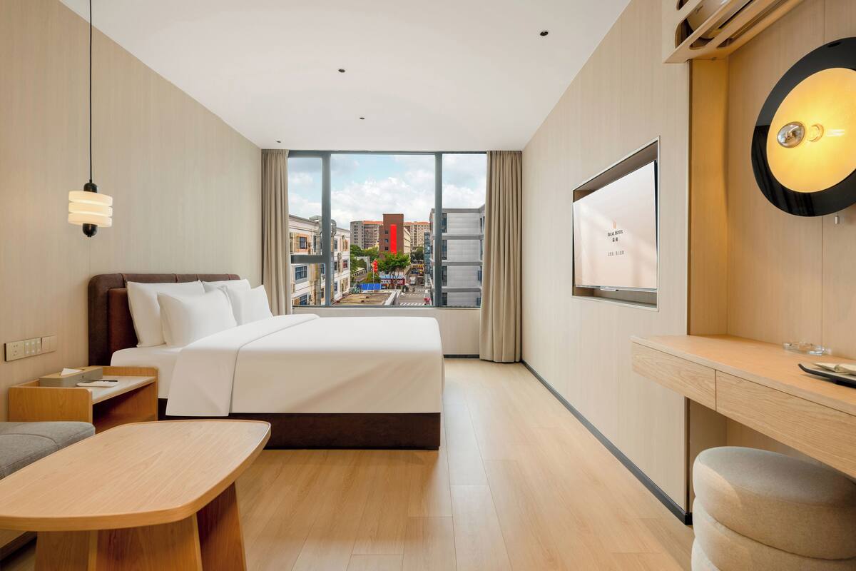 A modern hotel room with a large bed, a desk, and a view of the city through large windows.