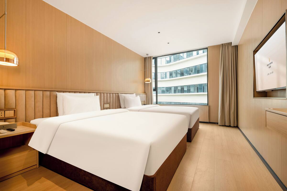 A hotel room with a large bed, a bedside table, a TV, and a window with a view of a building.