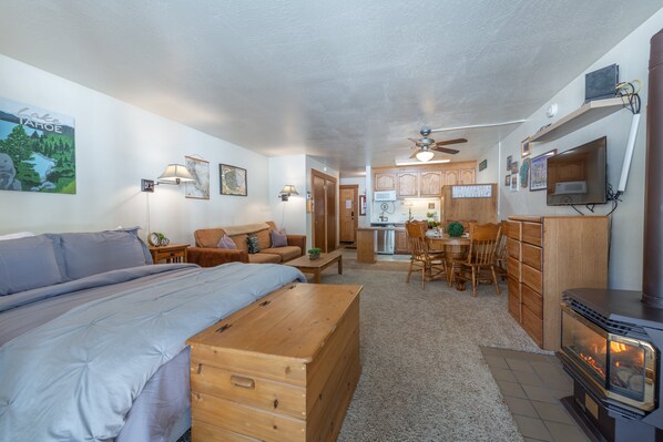 Un espacio habitable pequeño con una cama, un cofre de madera, una kitchenette y una zona de comedor.